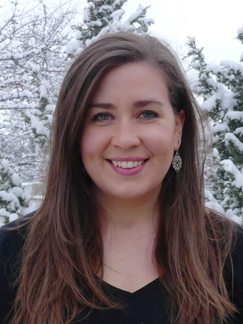 Lady with long brown hair and blue eyes with snowy trees behind her